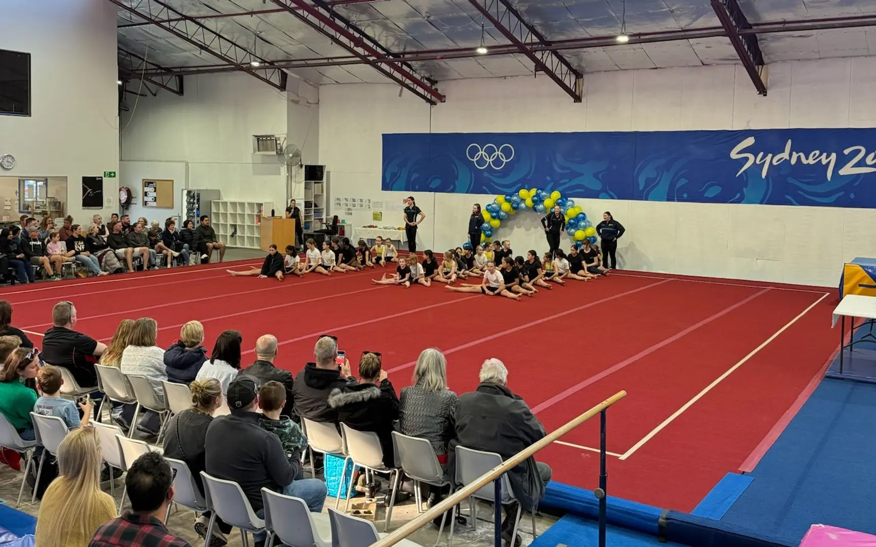 Families watching from the sidelines as gymnasts sit together on the competition floor beneath a blue and yellow balloon arch