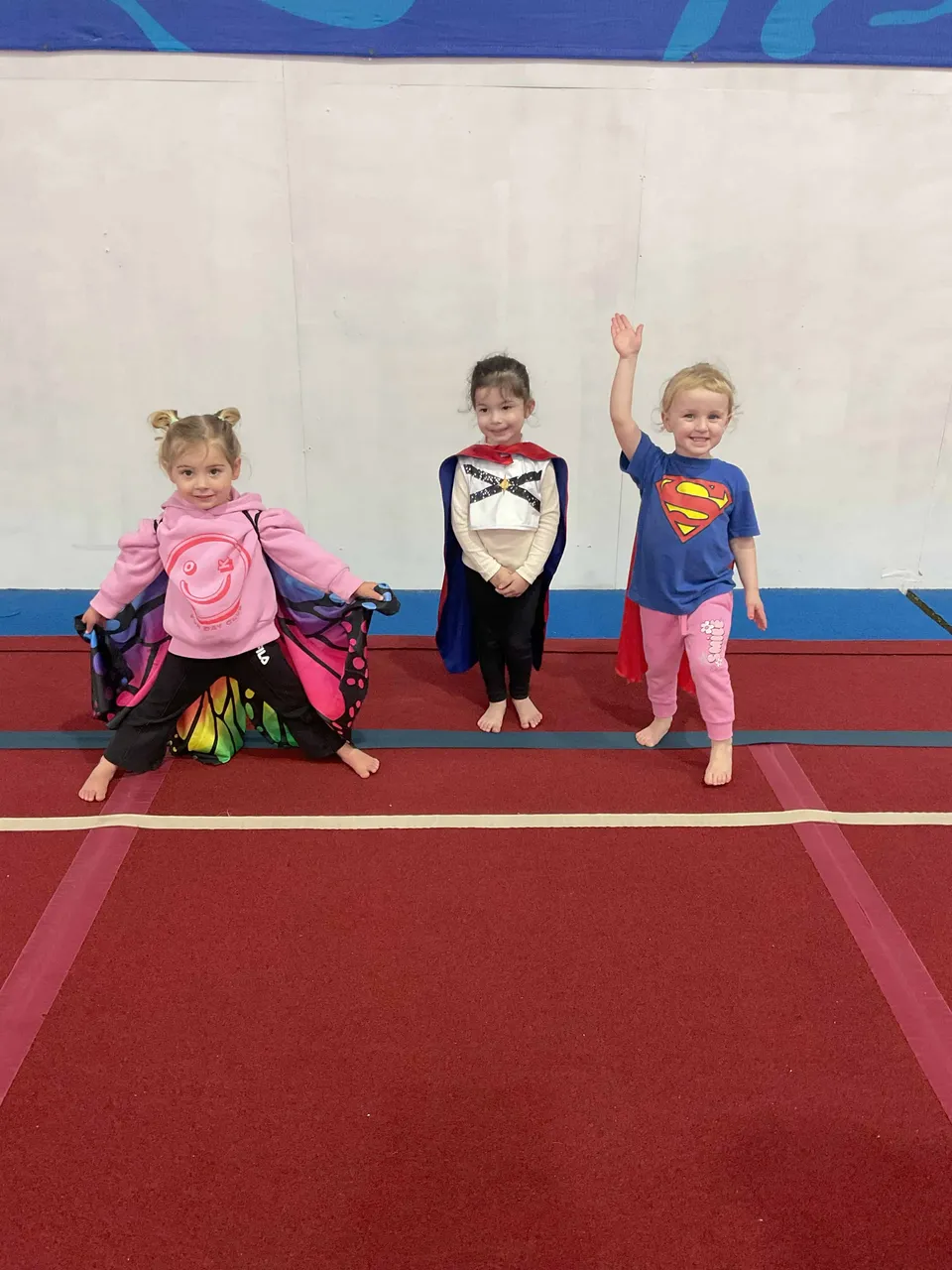 Three toddlers in superhero capes standing together on the gym floor